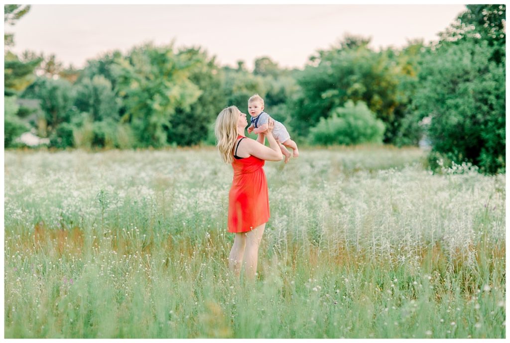 Eau Claire Family photos image taken in an open field Mom wears a red dress, holds baby up kisses his cheek sweetly