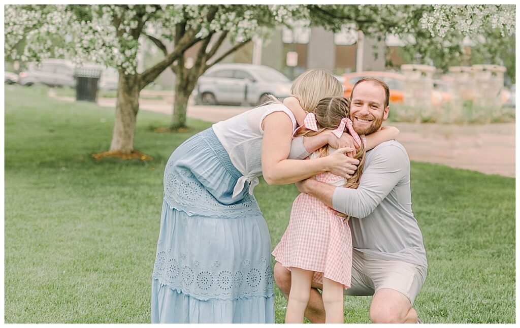 Parents wrap around around their daughter after she runs to them cheek while she laughs. Image is taken at a downtown park Side by side images of a family photo session. at Phoenix Park in Eau Claire, Wisconsin with blossoms in the background of the photo Wisconsin Wedding photographer. Eau Claire Family Photos.