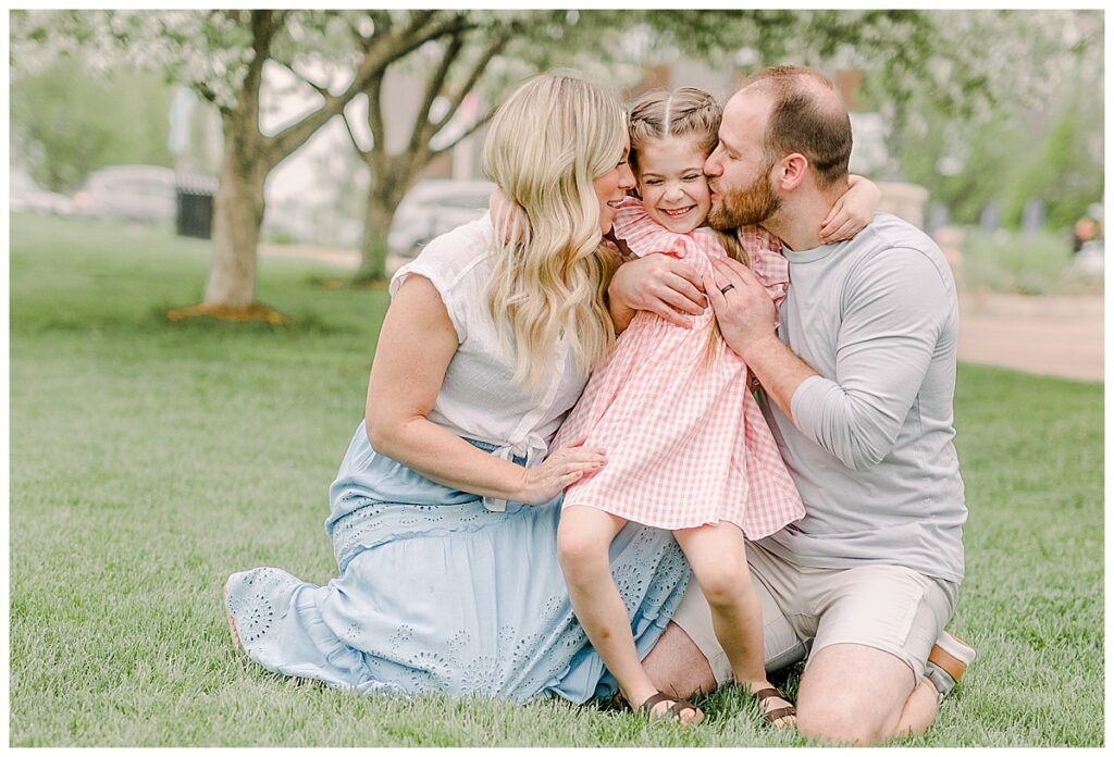 Parents snuggle their little girl who is wearing a pink dress. They each give her a kiss on the cheek while she laughs. Image is taken at a downtown park at Phoenix Park in Eau Claire, Wisconsin with blossoms in the background of the photo Wisconsin Wedding photographer. Eau Claire Family Photos.