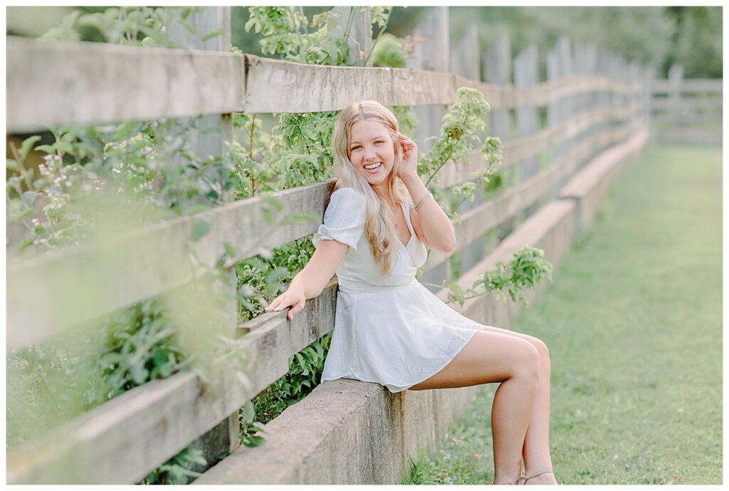 Senior poses for portraits  near menomenie Wisconsin. wearing  a white romper, leaning against a fence and laughing toward the camera