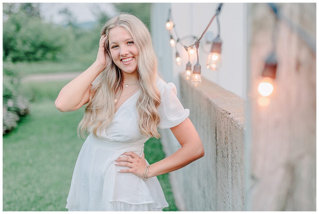 Senior poses for portraits  near Eau Claire Wisconsin. wearing a white romper, leaning agains a fence with twinkle lights