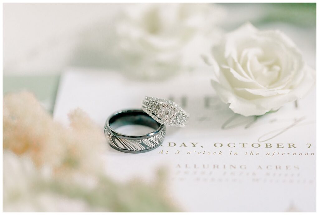 Image of the bride and groom's wedding ring, laid out with two white roses from the florists on top of it. The flat lay is set on top of the bride's wedding dress. Image by Alisha Marie Photography at the wedding venue alluring acres not far from Eau Claire, Wisconsin