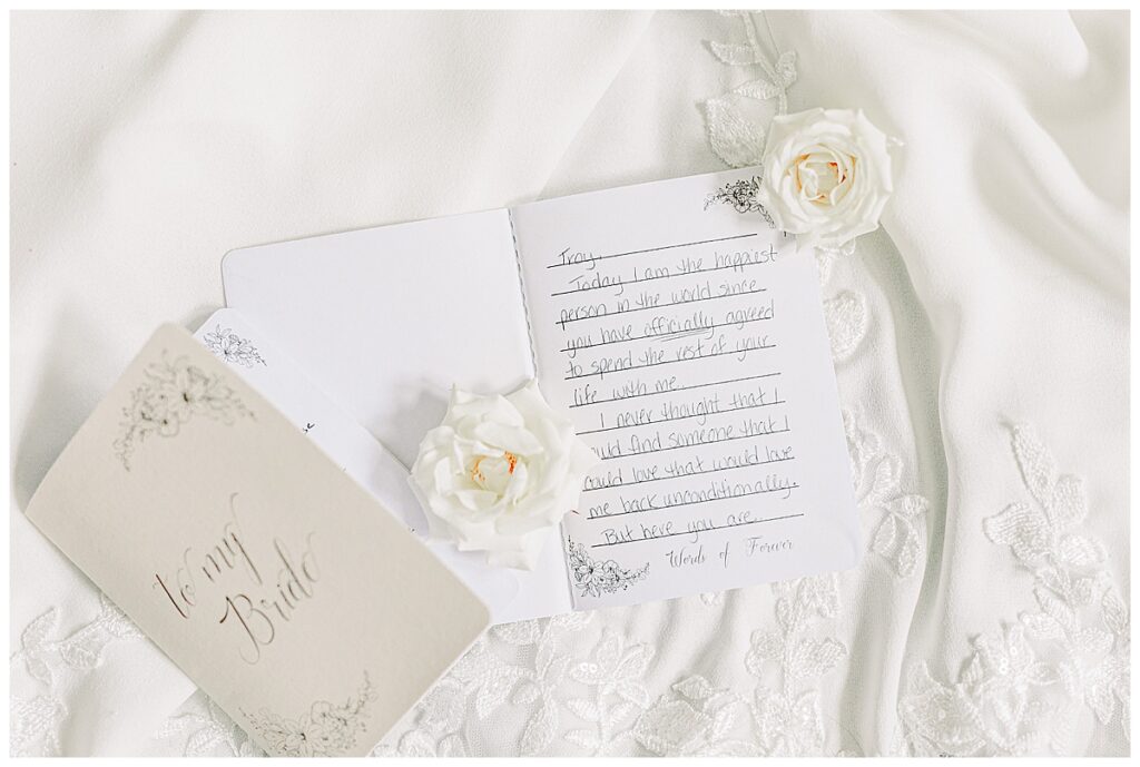 Image of the bride's private vow book, laid out with two white roses from the florists on top of it. The flat lay is set on top of the bride's wedding dress. Image by Alisha Marie Photography at the wedding venue alluring acres not far from Eau Claire, Wisconsin