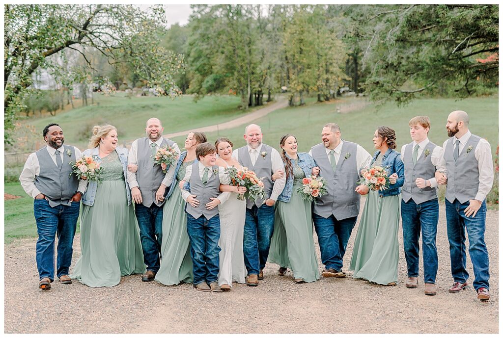 Wedding day wedding party portrait. Bridesmaids are laughing together. The women and bridesmaids wear a light green. The men wear jeans and a grey vest. Image by Alisha Marie Photography at the wedding venue alluring acres not far from Minneapolis, Minnesota and St. Paul