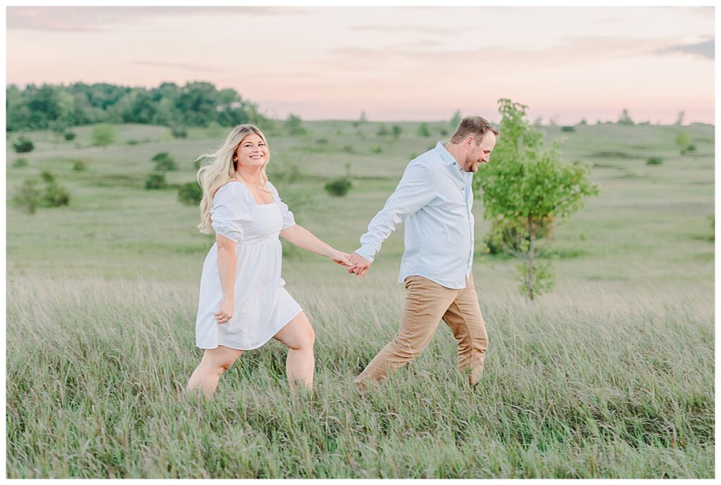 A wedding couple at their engagement session in an open field. she is wearing a white eyelet dress and he is wearing a light blue dress shirt. They are posing with a prompt from their photographer, Alisha Marie Photography, outside of Eau Claire, Wiscosin The woman laughs toward the camera while her fiancé walks hand in hand with her.  Alisha Marie Photography, outside of Minneapolis, Minnesota Image Taken By Eau Claire Wisconsin Wedding Photographer Family Photographer Senior Photographers